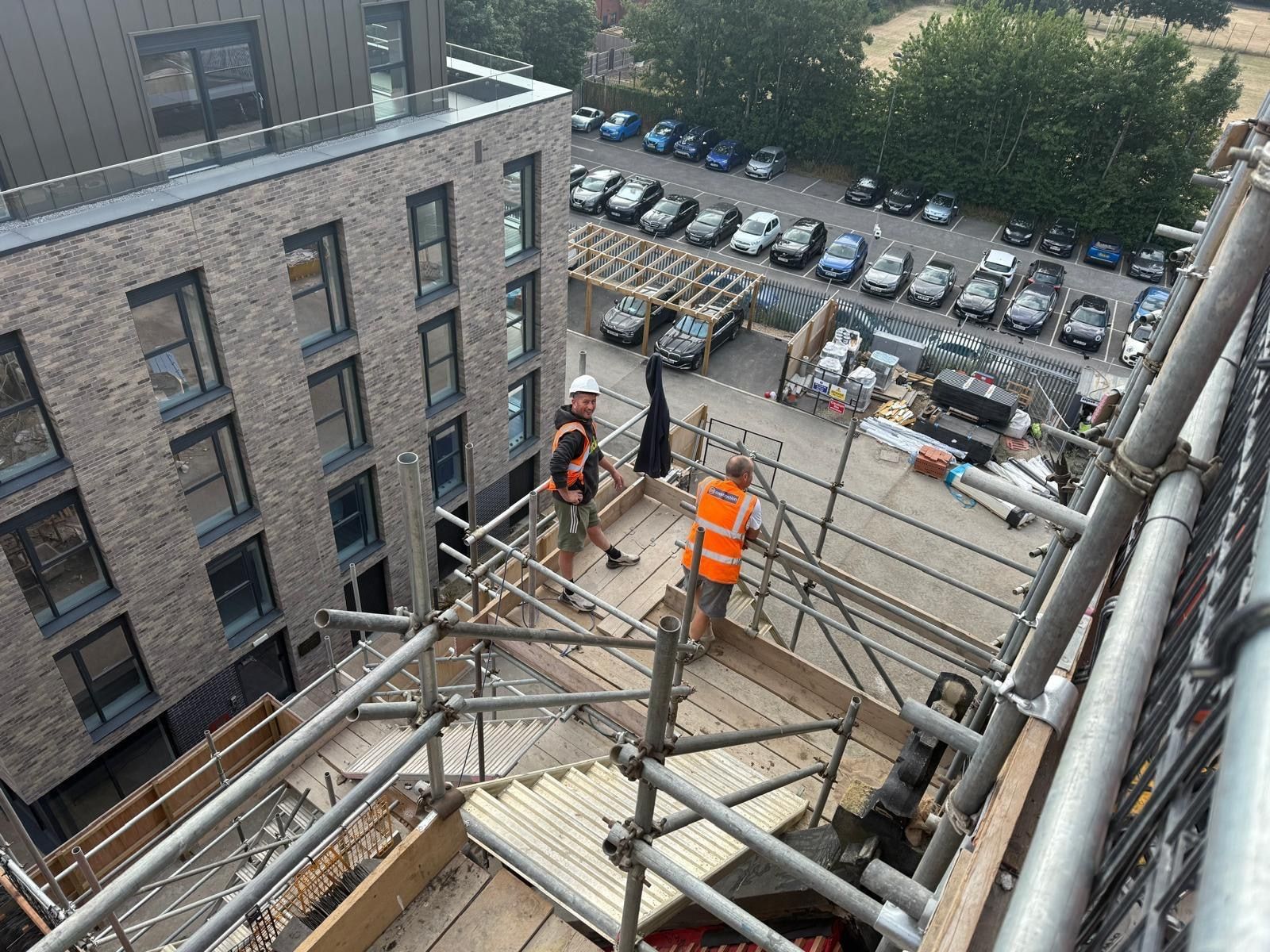 Two operatives in hi-vis on a scaffold platform high on a multi-storey residential development with brick and standing-seam cladding