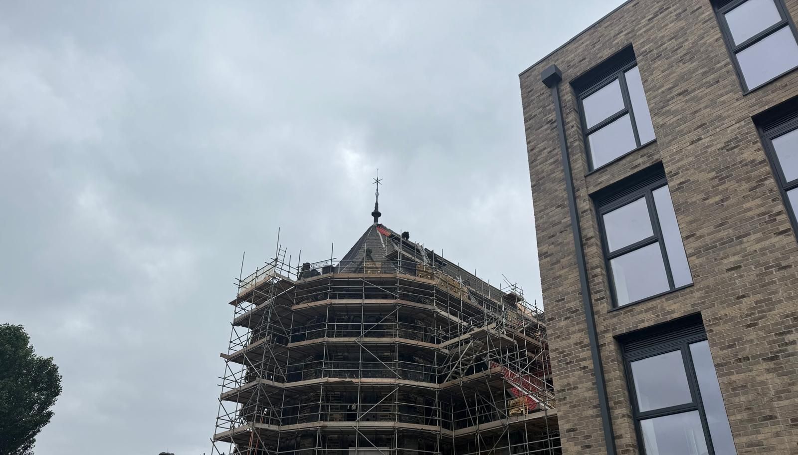 Large Victorian stone building wrapped in multi-lift scaffold to its spire, seen against a broad grey sky