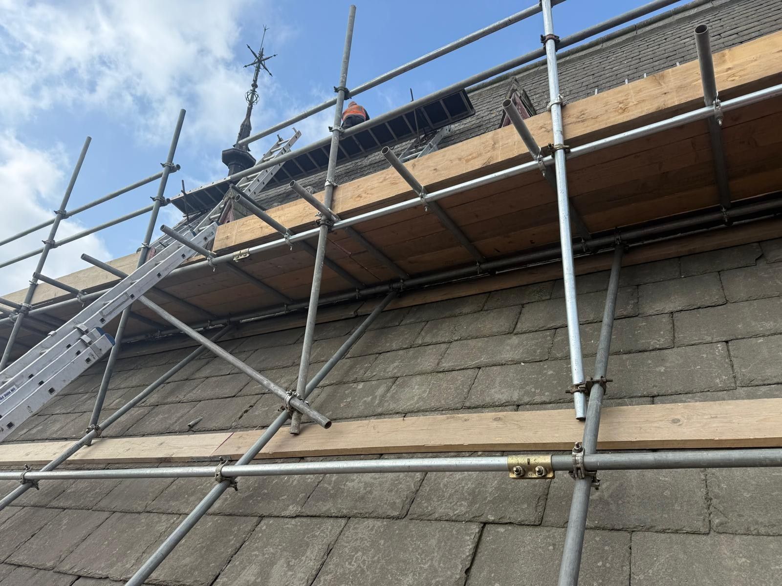 Scaffold lifts rising against a stone slate roof with a carved Gothic finial at the ridge