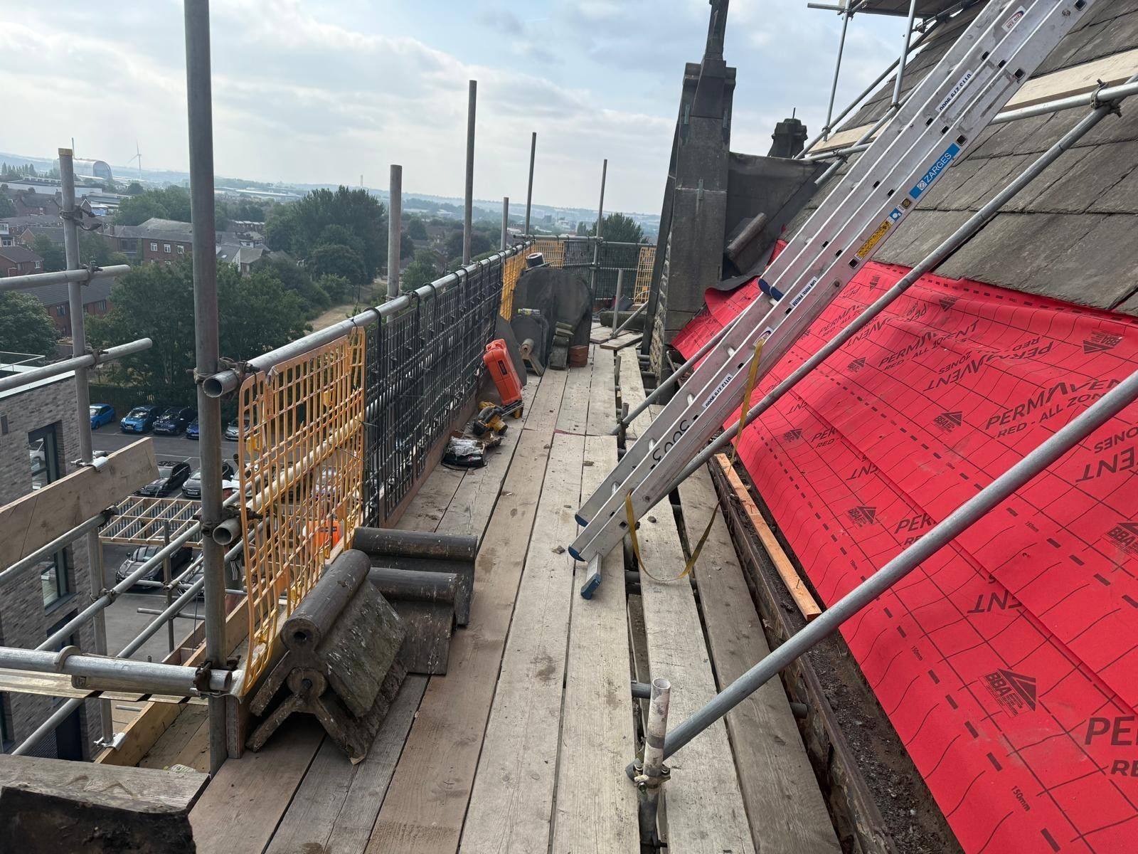 High-level scaffold run with stacked stone ridge pieces and a battened membrane slope, rooftops in the distance