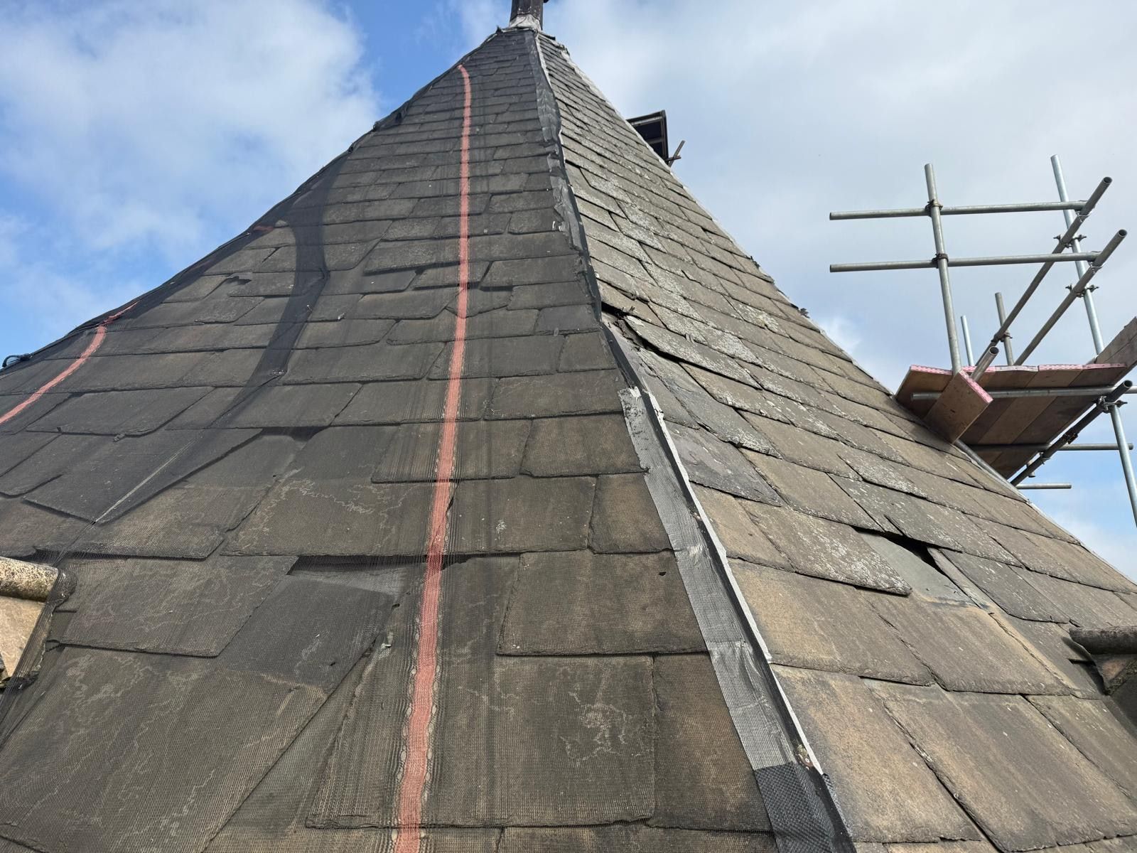 Steeply pitched heritage roof in weathered stone slate, one slope stripped to membrane with setting-out lines and a lead hip roll