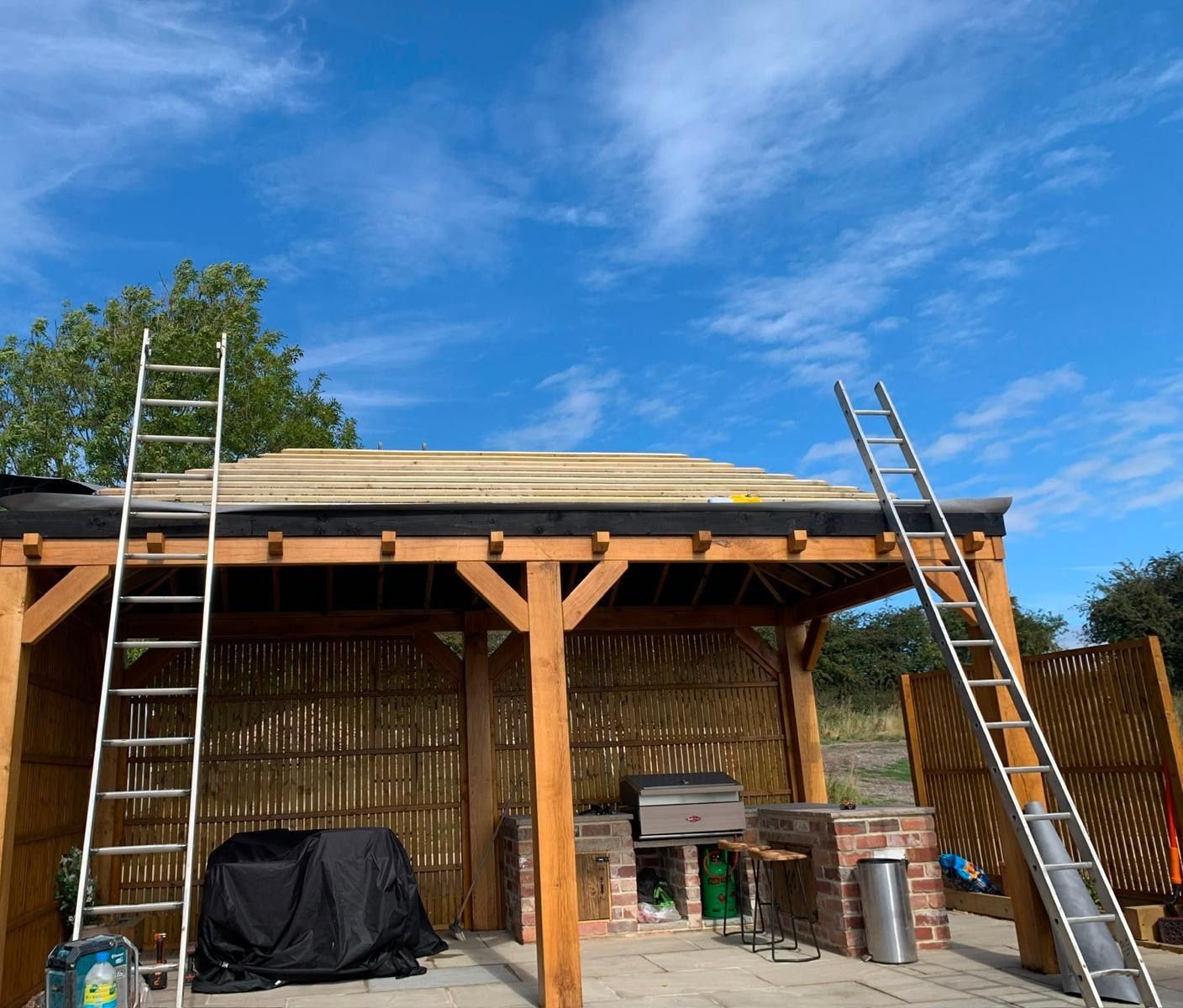Green oak frame of a garden building with roof deck boards being laid over the rafters, ladders in place