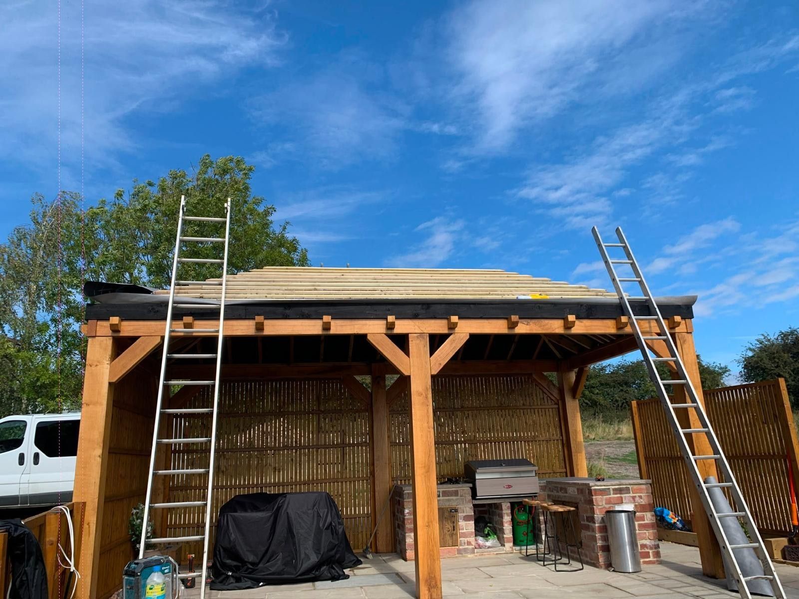 Green oak frame of a garden building with roof deck boards being laid over the rafters, ladders in place
