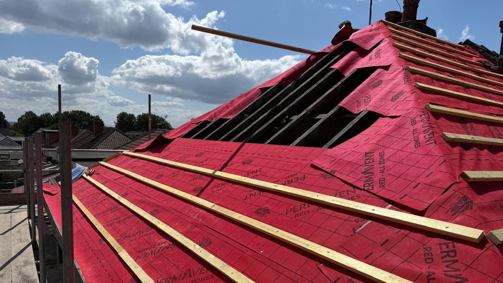 Re-roof in progress with red breathable membrane and new battens laid up to an open ridge, suburban rooftops beyond