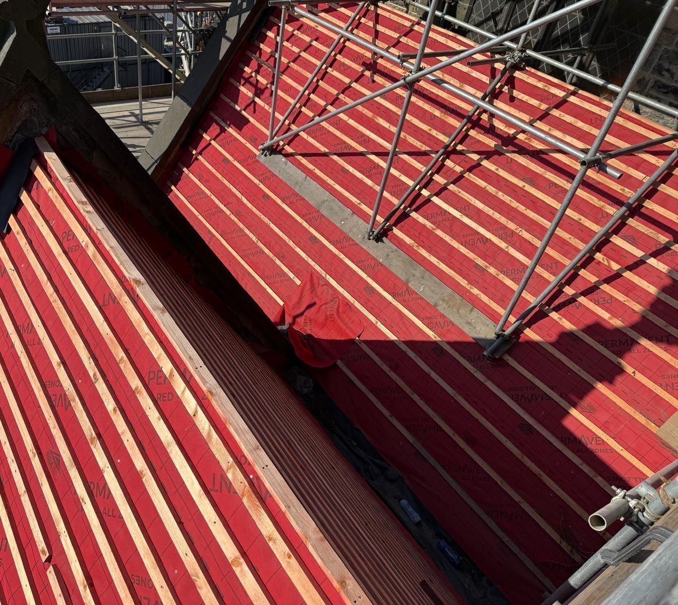 Roof valleys fully battened over red membrane in hard sunlight, with a lead-lined valley gutter and stone parapet