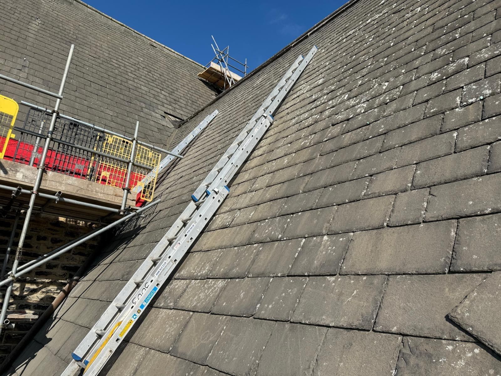 Steep weathered stone slate roof with roof ladders in place under a deep blue sky